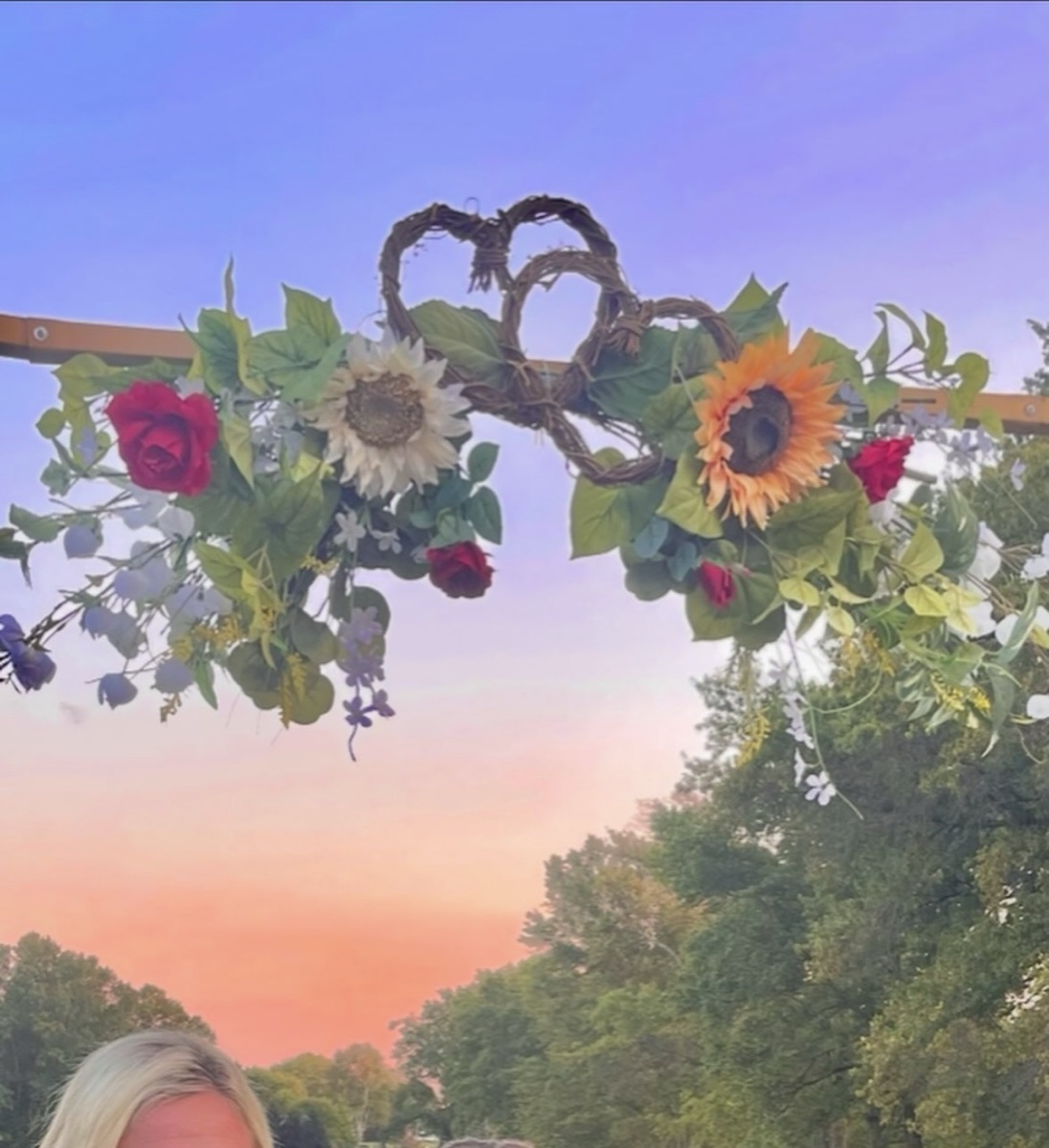 Outdoor floral arch with sunflowers, red roses and greenery against a sunset sky